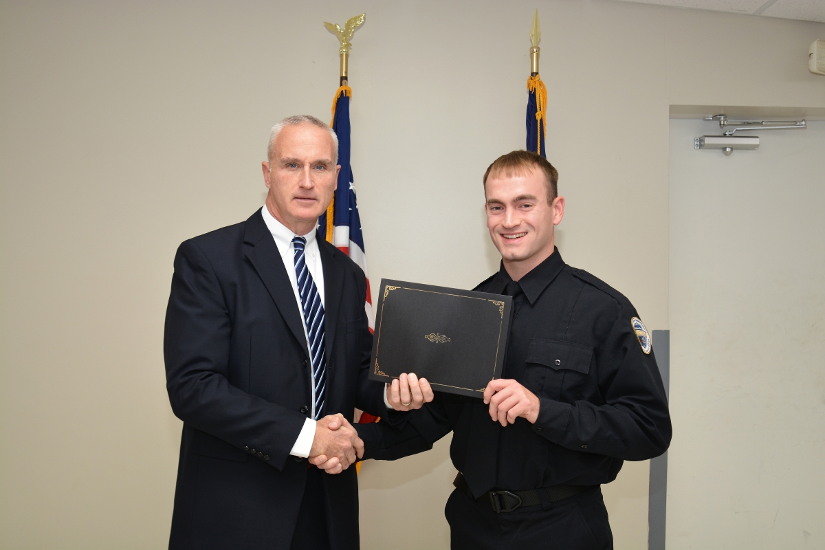 An instructor and a young man in a law enforcement uniform shake hands while posing with a diploma.