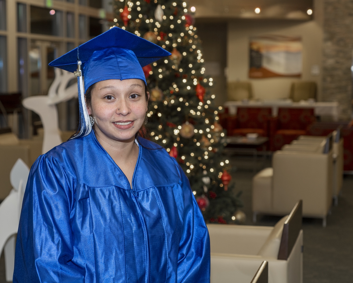 A young woman in a blue cap and gown smiles while sitting in front of a glowing Christmas tree.