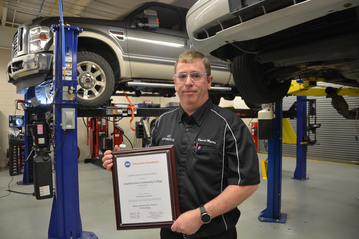 Man holds plaque in garage with cars on lifts in background.