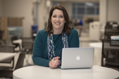 A woman smiles in a blue sweater while sitting behind a laptop