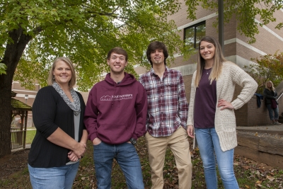Group of students stand outside underneath a tree on a fall day.