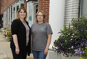 Two women stand together smiling on a sidewalk.