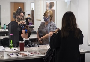 A young woman styles the hair of a mannequin while a female instructor watches