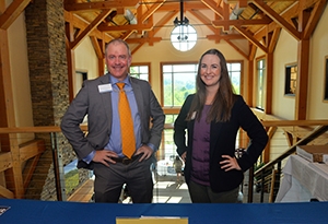 A man and woman dressed in suits smile while standing behind a table at a job fair.