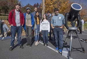 A group of students smile as they stand around a large telescope with their instructor