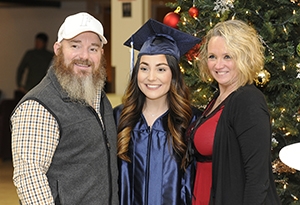 A mother and father smile while standing with their daughter wears a blue cap and gown at her graduation ceremony.