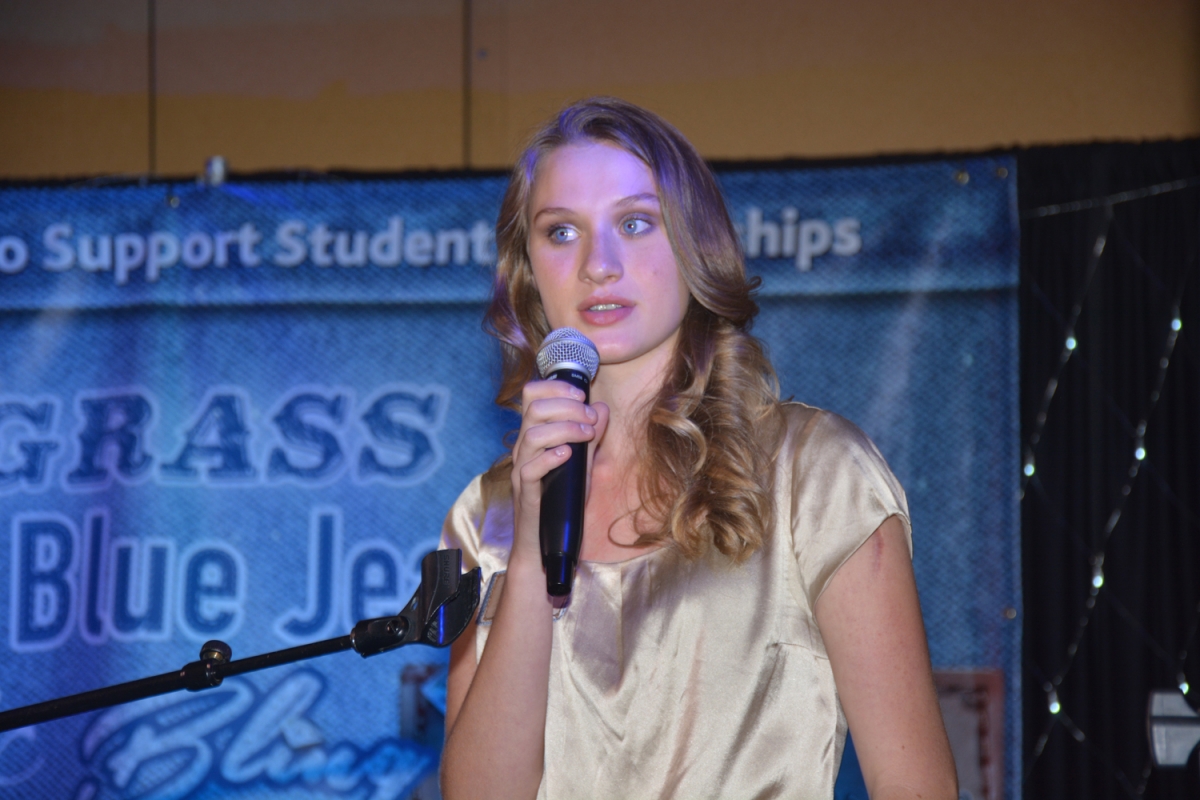 Young lady speaks into a microphone with a "Bluegrass, Blue Jeans & Bling" banner behind her.