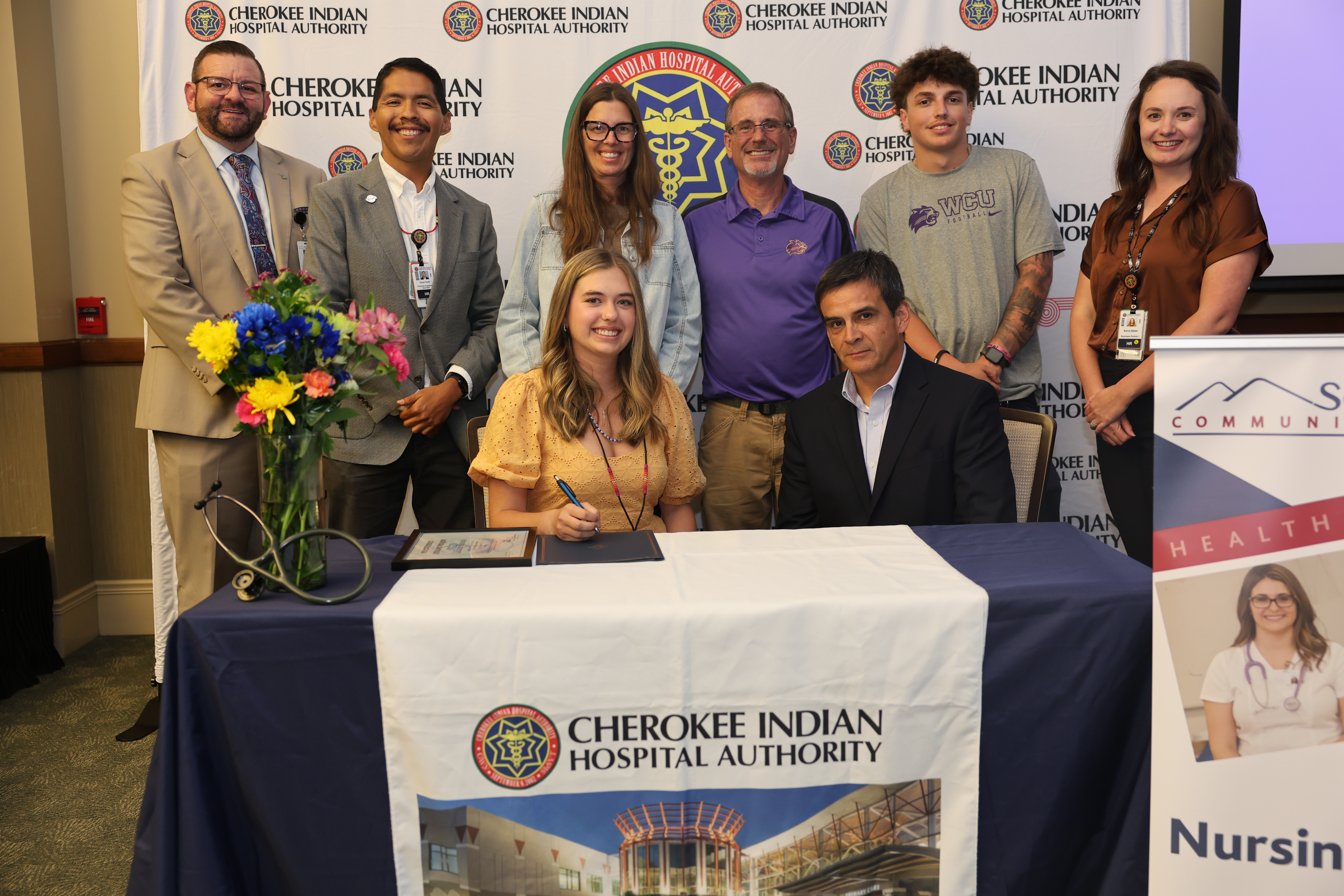 Student signs paper surrounded by family and college and hospital officials
