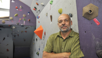 man stands in foreground; indoor rock climbing wall is behind him.
