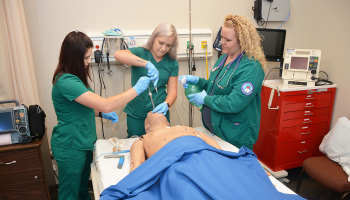 Three female students wearing turquoise scrubs practice their respiratory therapy skills on a simulation mannequin inside a room on SCC's Jackson Campus in Sylva.