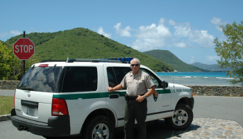 Man wearing NPS uniform stands beside law enforcement vehicle near scenic beach.