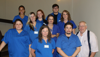 Ten students wearing blue scrubs stand with their professor on a stairway inside a building on SCC's Jackson Campus.