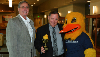 Two men stand beside a life-sized duck mascot. One man holds a duck-topped trophy.