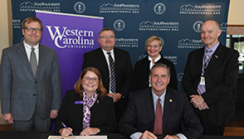 A group of academic leaders gather around in suits and smile as they sign documents