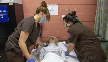 Two females in brown scrubs demonstrate nursing skills on a mannequin