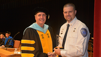 A male college president in a cap and gown presents an award to a male graduate from the emergency medical science program