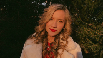 A young woman with blonde hair and red lipstick stands outside for a picture