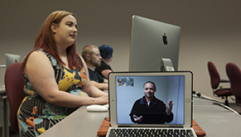A young woman is working on a computer while an instructor is video chatting the class from an ipad