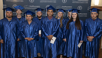 A group of students in blue caps and gowns smile as they sit and wait to receive their diplomas.