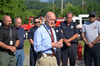 Curtis Dowdle speaks; first responders and emergency vehicles are in the background.