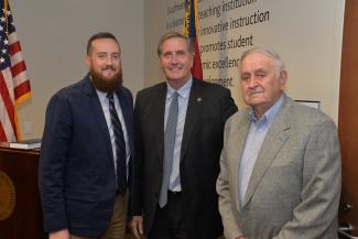 Three men pose in a board room in front of U.S. and N.C. flags.