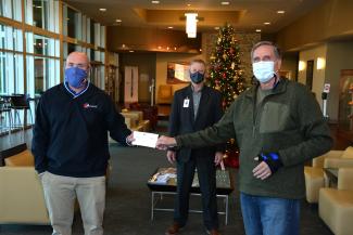 Two men exchange a check while a third stands behind them. All three are social distancing and wearing masks.