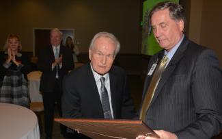 Two men look at a plaque as people in the audience applaud.