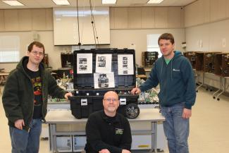Jim Falbo kneels and two students stand in front of the SWAT robot case