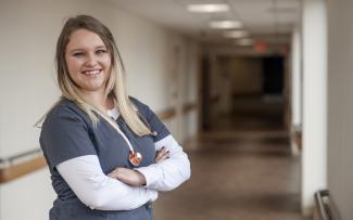 Nurse wearing scrubs and stethoscope stands in hospital hallway.