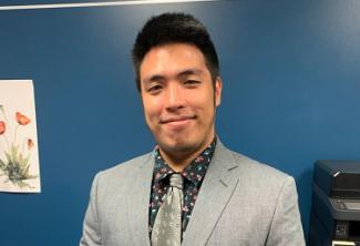 A young man smiles in a gray suit with a floral shirt and silver tie