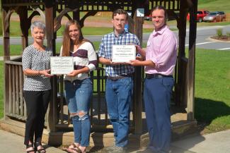 Two young students are holding scholarship certificates in front of a gazebo