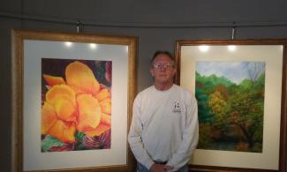 Randy Lanier standing between two paintings one is a flower the other a tree scene
