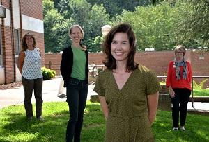 A group of four women stand safely apart while smiling outside