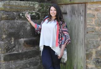 A young woman stands outside of an old wooden building surrounded by nature.